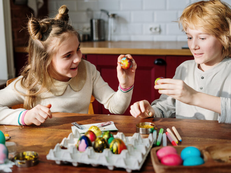 a young brother and sister use pens, paints and glitter to decorate eggs
